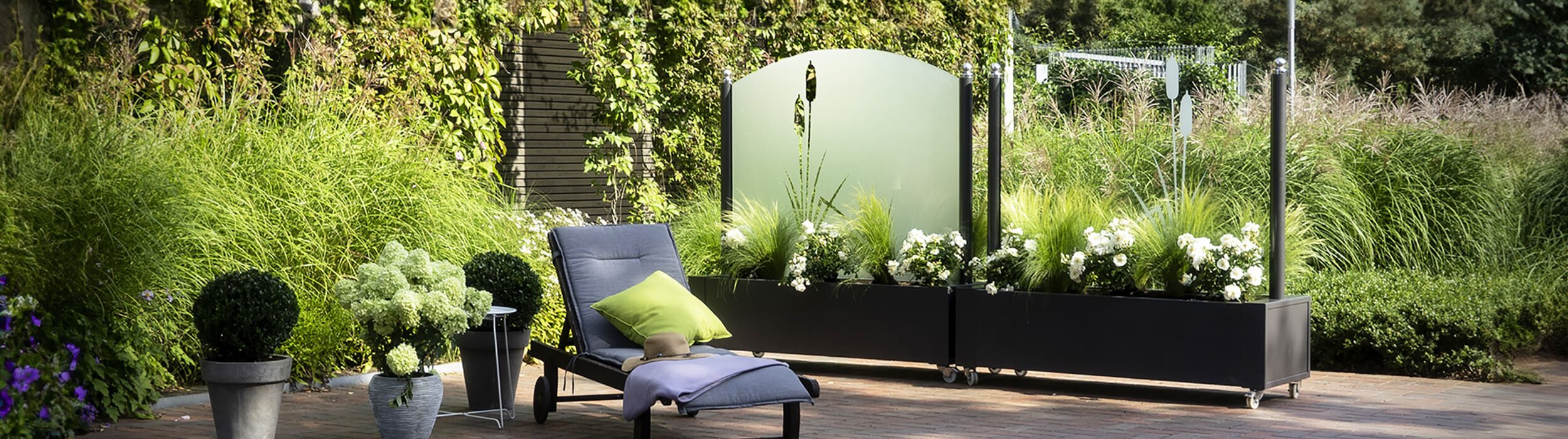 A lounge chair, three plants, and two wind protection elements on a terrace.
